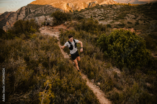 Bilde på lerret Endurance athlete running along a narrow trail through rugged mountain terrain surrounded by wild vegetation and rocky landscape