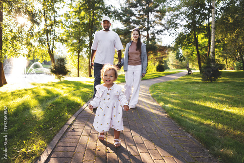 Happy family with small child walking together in green park at sunset. Warm summer outdoors with golden sunlight. Family lifestyle and parenting in nature