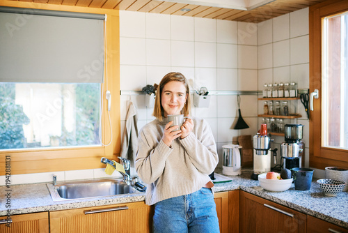 Young woman standing in cozy home kitchen holding warm coffee mug and smiling near window in morning light relaxed lifestyle moment with simple home interior and daily routine