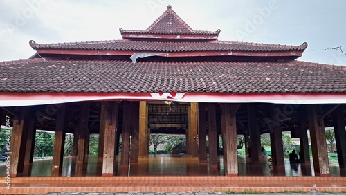 Javanese Pendopo traditional wooden pavilion architecture, frontal view of historic Indonesian Joglo building with tiered clay tile roof and red white decorative banners in cultural site.