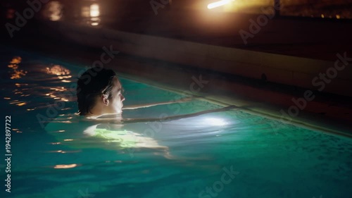 A serene video shows a teenager relaxing in an illuminated swimming pool at night. Underwater lights cast a tranquil green glow, highlighting subtle movements and reflections