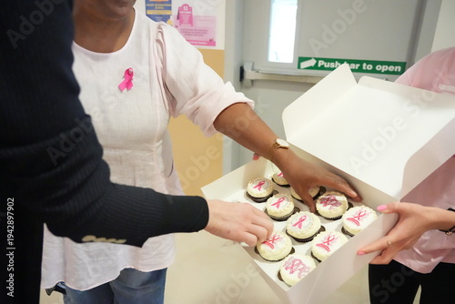 Participants from diverse backgrounds take cupcakes decorated with pink ribbon symbols during a breast cancer awareness event in a school hallway.