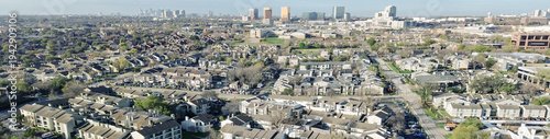 Panorama aerial urban edge overlook near Fair Oaks Ave and Walnut Hill Ln reveals apartments bordering wooded trails with skyline rising beyond. Sloped roofs, mixed facades, cool winter, Dallas