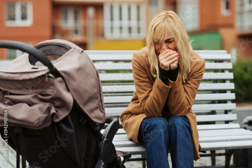 Worried young mother sitting on a bench next to a stroller in an urban park