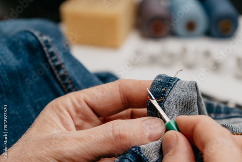 tailor is using green seam ripper to take apart stitches on denim fabric. scissors and sewing supplies are present on table. setting shows crafting area with tools. closeup.