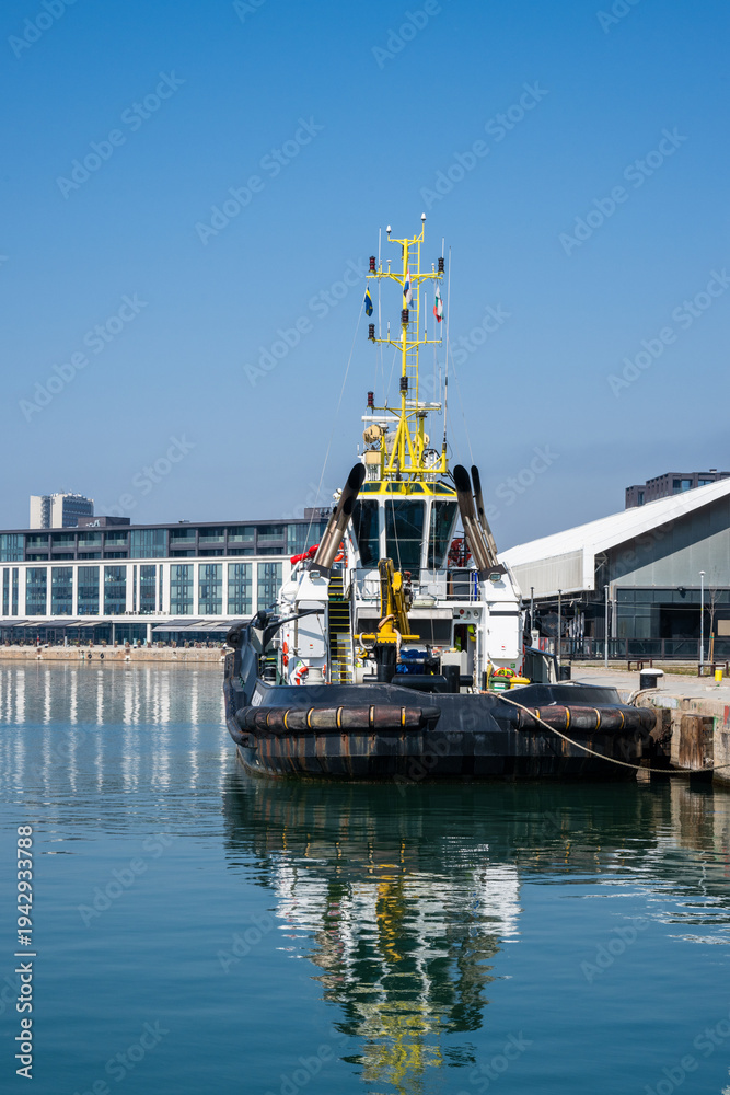 Fototapeta premium a black, white, and yellow tugboat moored at a concrete pier, reflecting perfectly in the calm harbor water, with modern waterfront buildings in the background