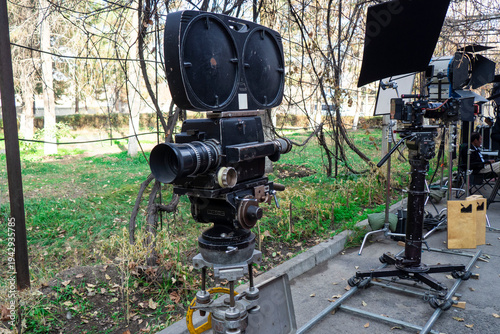 Old professional cinema camera with interchangeable lenses mounted on tripod near dolly rails during film shooting. Retro analog filmmaking equipment on movie production set.