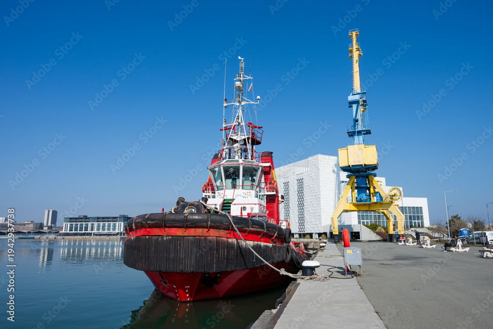 Fototapeta premium a red and black tugboat moored at a concrete dock next to a massive yellow and blue portal crane, with a modern white building and the city skyline