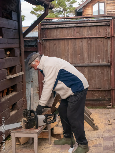 Lumberjack cutting wooden logs with electric chainsaw in backyard. Caucasian man preparing firewood for heating house. Seasonal rural work, manual labor and energy supply concept.
