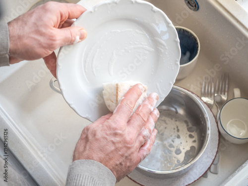 Man washes dishes in a kitchen sink with a sponge and liquid soap under running water. Contemporary domestic scene and modern masculinity. Concept of sharing household chores. Day light, close-up