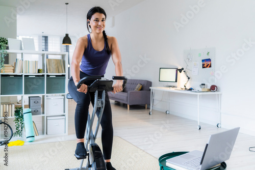 Smiling young woman listening music through bluetooth while sitting on exercise bike at home