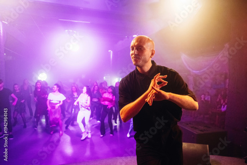 Male dance instructor demonstrates choreography in a vibrant dance studio with colorful lighting and a group of students practicing in the background