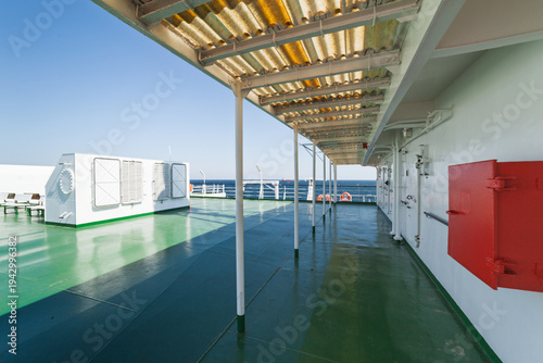 Spacious upper deck of ferry vessel with green floor and white metal ventilation units. Distant sea horizon visible over railing under blue sky. Red fire equipment box stands on wall.