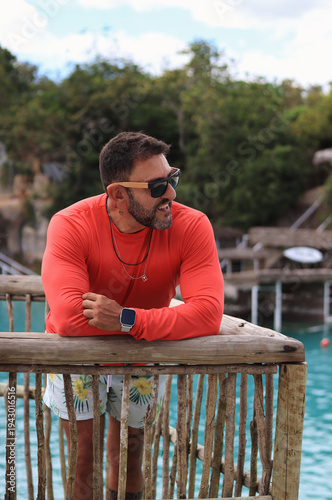 Smiling man in red rash guard and sunglasses leaning on rustic wooden railing overlooking turquoise water