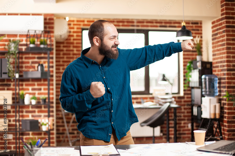 Fototapeta premium Caucasian male manager dancing with excitement in startup office after securing business deal. Young man with beard punching the air with joy after successfully completing project presentation.