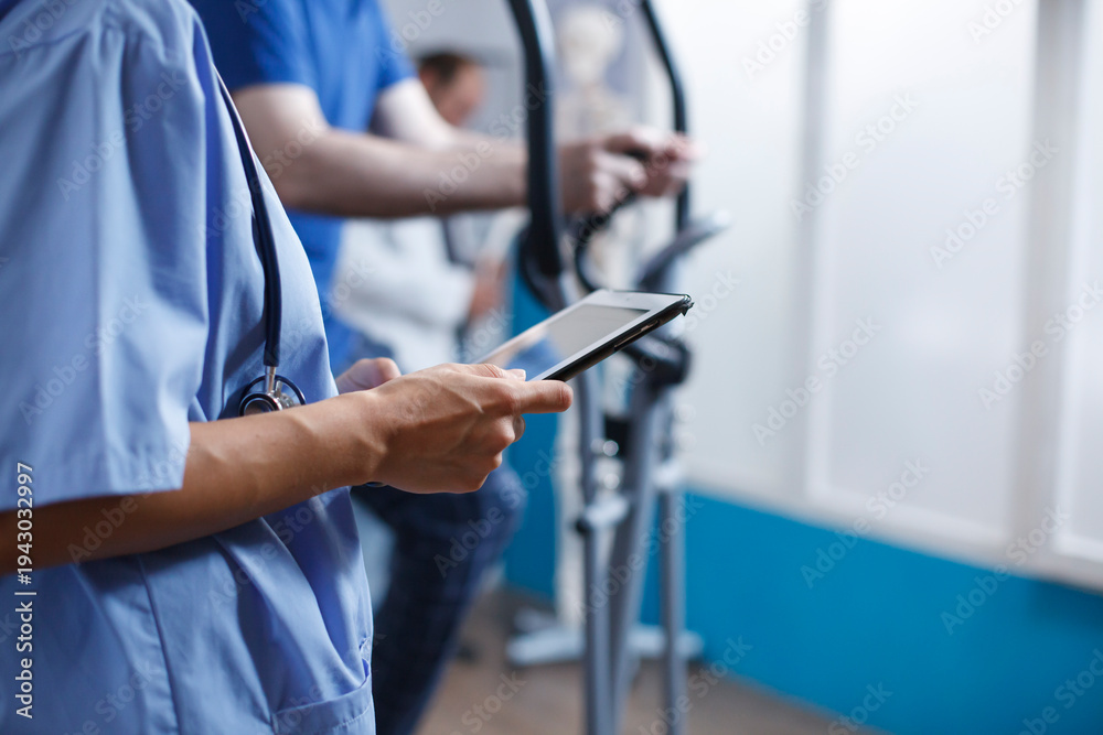 Obraz premium Close-up of nurse using a tablet at clinic, reading a patient report while wearing blue scrubs and stethoscope. Caucasian female physician holding digital device for medical research.