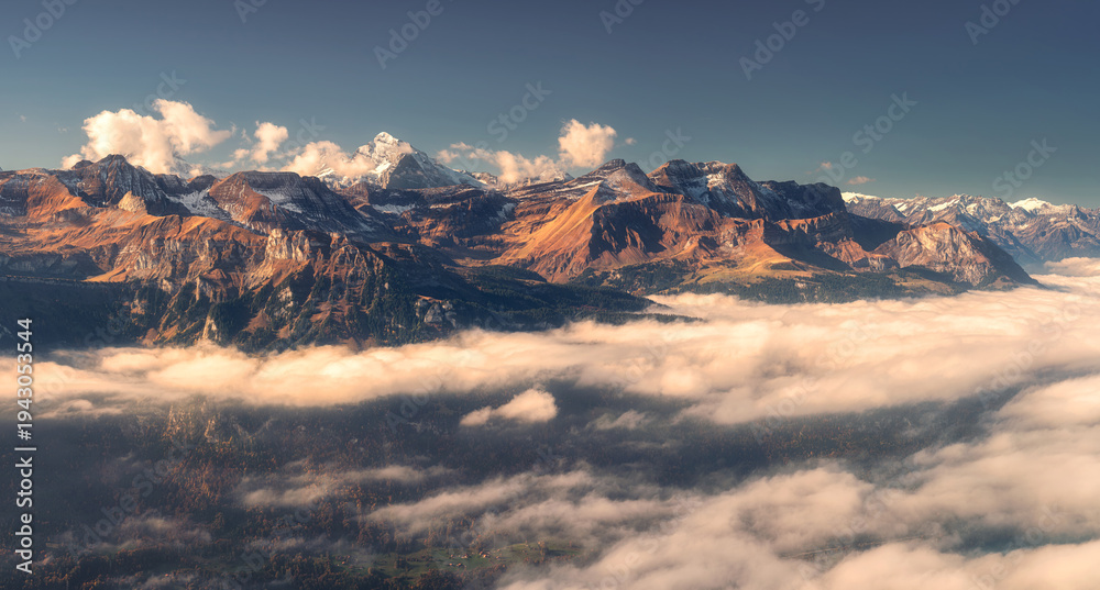Fototapeta premium Panoramic aerial view of the Swiss Alps with snow-capped peaks and low clouds at sunset. Bright evening light and clear blue sky over rugged alpine mountains in Switzerland. Top view. Landscape