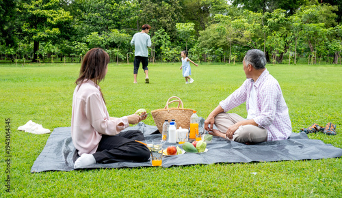 Obraz na plátne Mother and grandfather sit on mat in grassfield of public park and look father and grandchild enjoy to play together with daylight