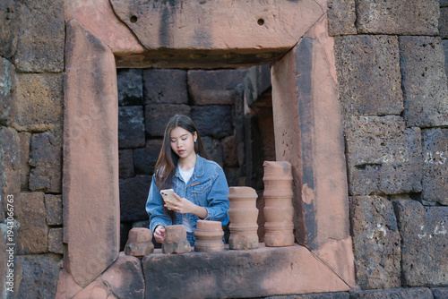 A young woman stands at an ancient stone window, using her smartphone while exploring historic temple ruins.