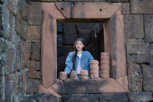 A young woman stands by an ancient stone window surrounded by historic temple ruins and clay artifacts. The scene captures a calm moment of exploration.