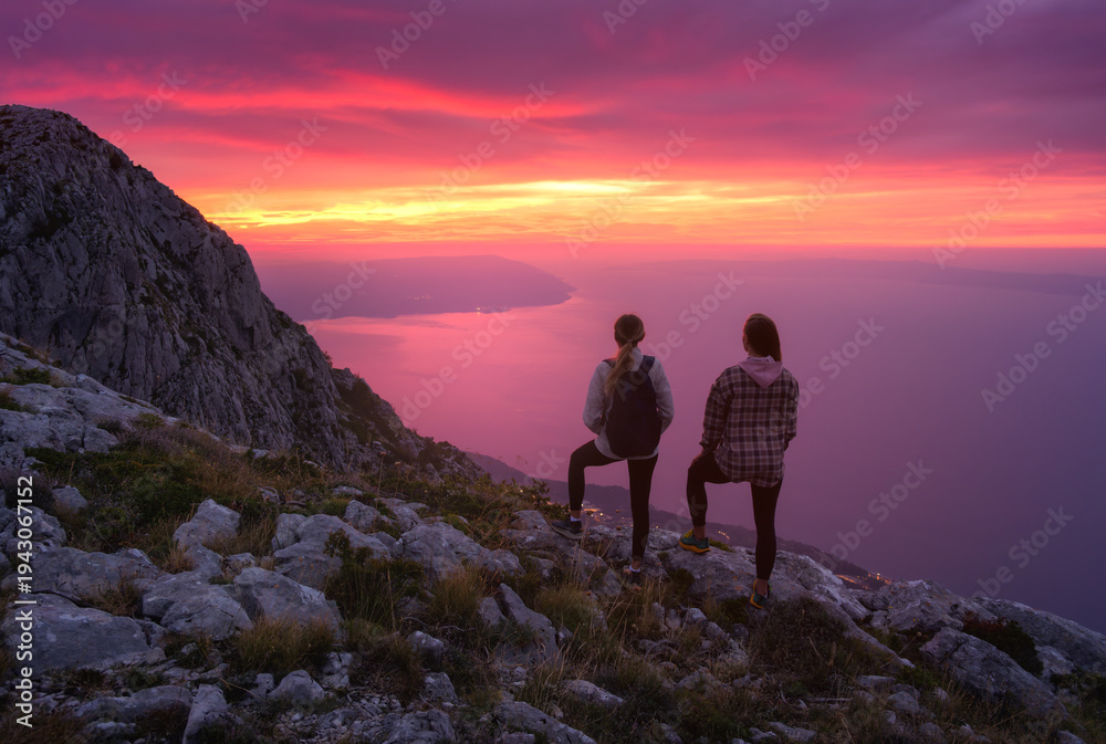 Obraz premium Two women hikers with backpacks standing on rocky mountain peak at sunset, overlooking the sea with colorful pink sky. Outdoor travel adventure. Silhouette of girls on mountain at dusk. Lifestyle