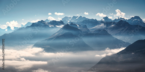 Panoramic aerial view of the Swiss Alps with snow-capped peaks and low clouds at sunrise. Bright morning light and blue sky over rugged alpine mountains in Switzerland at dawn. Top view. Landscape