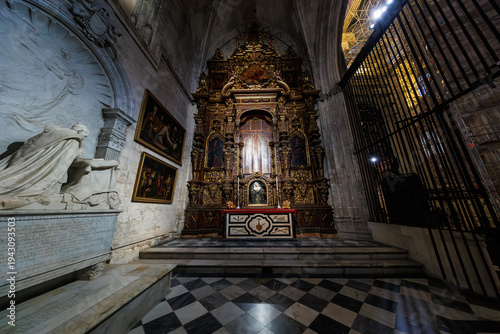 Bild auf Leinwand Baroque altarpiece with gilded wood, paintings, and reliquary stands in a side chapel in Seville, Spain, beside a white marble tomb and checkerboard stone floor