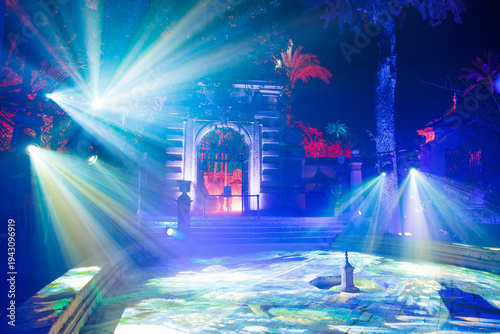 Seville, Spain. Night courtyard at the Real Alcazar shows a baroque stone arch and gate lit by blue, violet, and red lights, foggy rays, tiles, fountain, and palms.