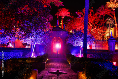 Night walkway in Real Alcazar gardens, Seville, with magenta and cobalt lighting casts palm and orange trees as a bright orb shines from the arched gate at center.