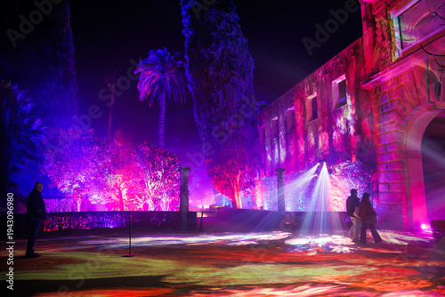 Night light festival in Real Alcazar gardens, Seville. Magenta, violet, and blue projections wash stone walls as visitors wander past columns and an arched portal.