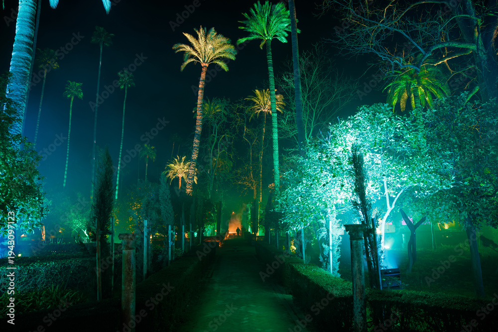 Fototapeta premium A nocturnal walkway in the Real Alcazar gardens in Seville shows turquoise and emerald lighting, tall palm silhouettes, fog, stone posts, and a lone person at a vanishing point.