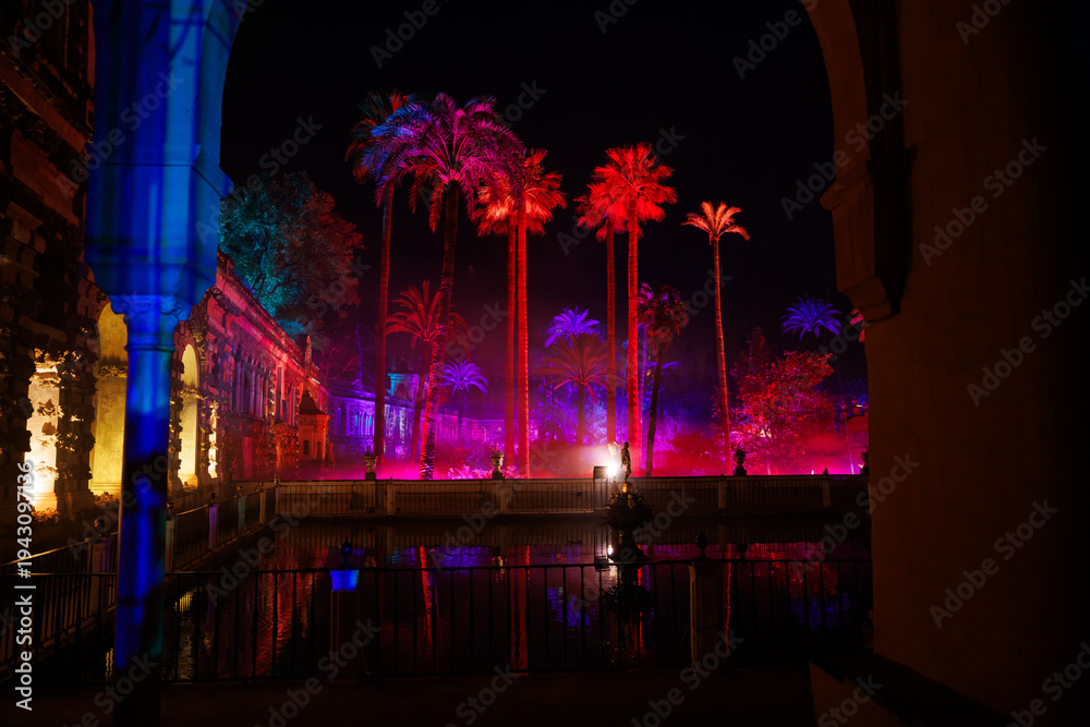 Obraz premium Nighttime light spectacle at the Real Alcazar gardens in Seville, where towering palms glow above a reflective pool, framed by Moorish arches and stone arcades.
