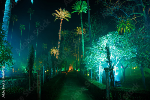 A nocturnal walkway in the Real Alcazar gardens in Seville shows turquoise and emerald lighting, tall palm silhouettes, fog, stone posts, and a lone person at a vanishing point.