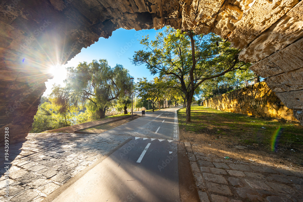 Fototapeta premium Seville, Spain, a sunlit path under a stone arch with a bike lane leads by a golden wall and trees. Morning light casts long shadows. A trash bin and lamppost stand nearby.