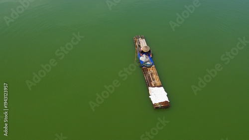 фотографія Fisherman casts net over calm on a bamboo raft, greenish water