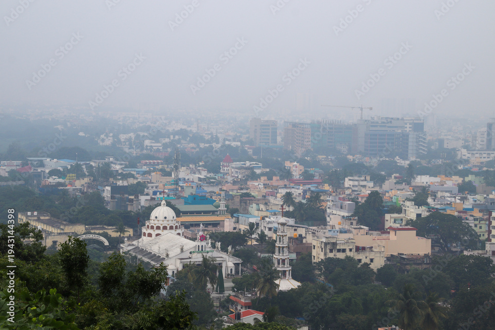 Obraz premium A hazy cityscape with dense buildings and a prominent church dome viewed from a green hillside in Chennai City 