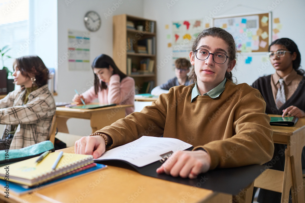 Obraz premium Portrait of Caucasian teenage boy with long hair sitting at desk in classroom looking into camera, while other multiethnic teenagers studying in background during lesson
