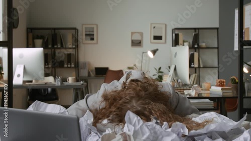 Frustrated businesswoman putting her head down on desk, cluttered with piles of crumpled documents in office