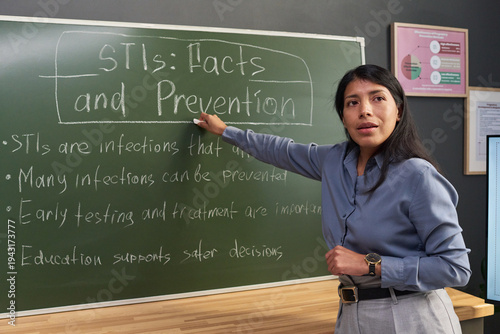 Hispanic woman standing in classroom pointing at chalkboard teaching about sexually transmitted infections prevention, educational materials visible in background