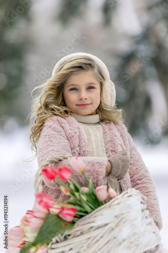 A beautiful little girl in pink clothes with a basket of flowers walks along a path in a snowy forest. Spring festival