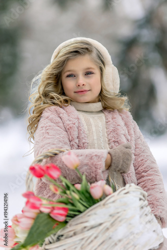 A beautiful little girl in pink clothes with a basket of flowers walks along a path in a snowy forest. Spring festival