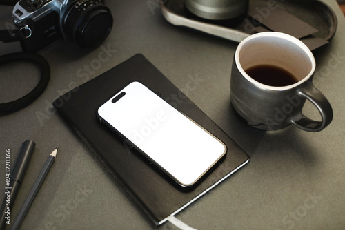 Close-up of a mobile phone with a blank screen on a notebook with a cup of black coffee and camera on a desk