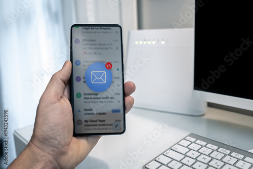 Closeup of male hand holding smartphone displaying email inbox interface with red notification bubble icon of unread messages in white home office with computer keyboard and wifi internet router