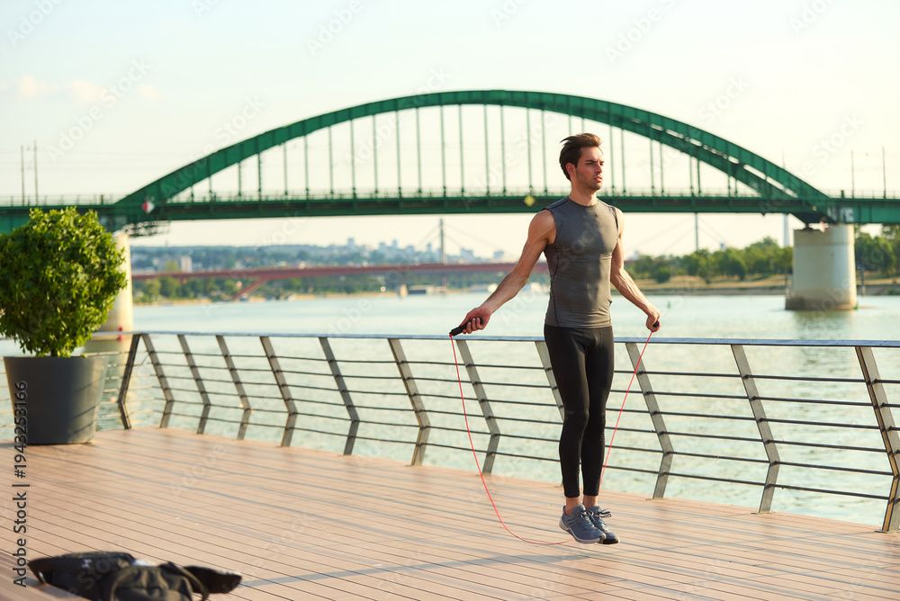 Obraz premium Male athlete skipping rope for fitness and cardio workout on a riverside pier with a green arch bridge spanning the river in a city during daylight