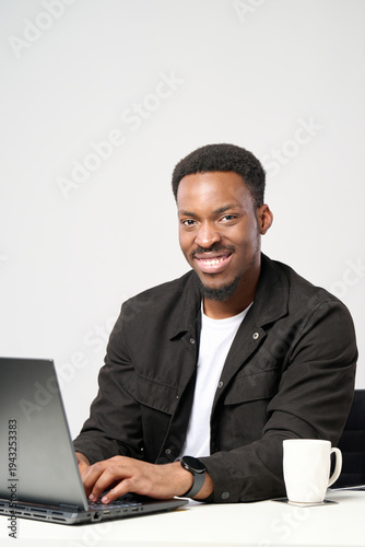 Young african american man working remotely on a laptop, smiling confidently and happily, embracing professional development and modern technology in a bright workplace