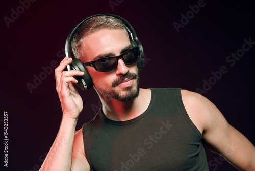 Fashionable man wearing headphones and sunglasses, adjusting one ear cup while listening to music with a confident expression under moody red illumination