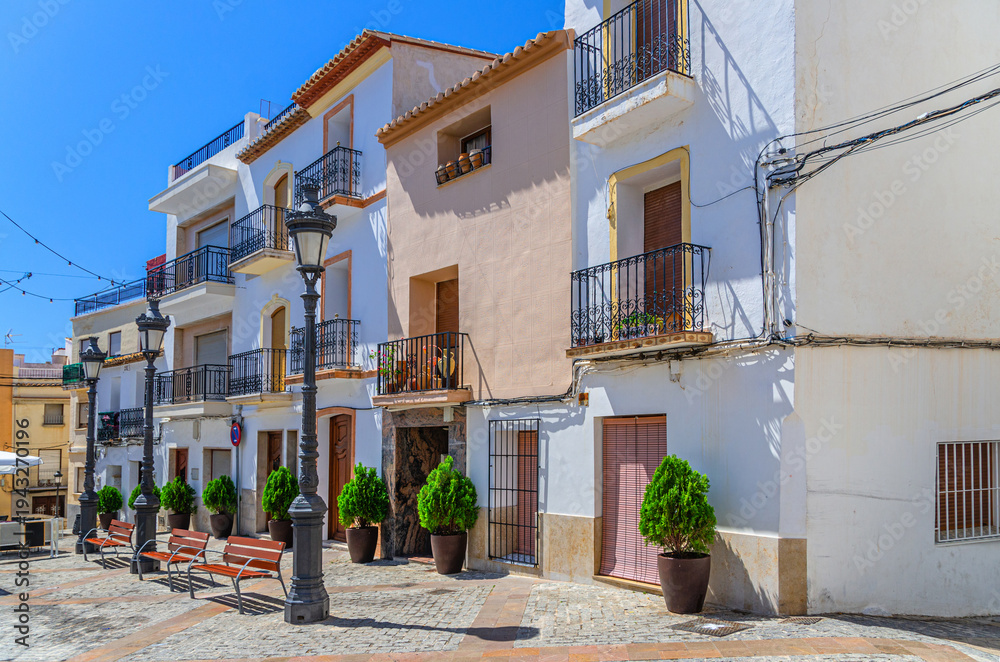 Obraz premium Old town Calpe city historic centre with white houses traditional buildings, benches and street lights on small square Villa Plaza in sunny summer day, Calp Casco Antiguo, Valencian Community, Spain