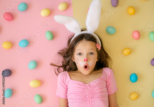 Cute surprised girl with bunny ears and face paint lying among colorful Easter eggs on pastel background. Easter holiday, childhood fun and festive spring concept.