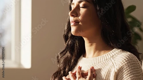 Woman meditating in soft natural light. Hands over heart with eyes closed in quiet reflection. Mindfulness and self-care concept