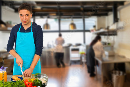 Young man wearing a bright blue apron holds a plate of cookies and drinks a glass of milk in a warm kitchen setting, suggesting homemade baking, casual hospitality and a tasty snack moment.
